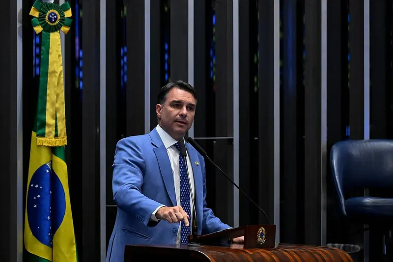 Flávio Bolsonaro, de terno azul-claro e gravata azul-marinho, fala ao microfone da tribuna do Senado, ao lado da bandeira do Brasil