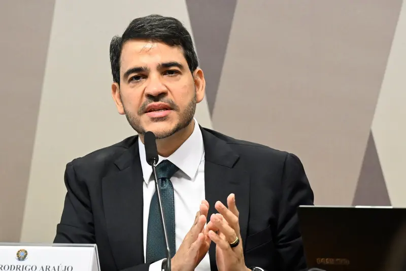 Jorge Messias, de barba, terno escuro e gravata verde, fala diante de um microfone, com as mãos unidas à frente do peito