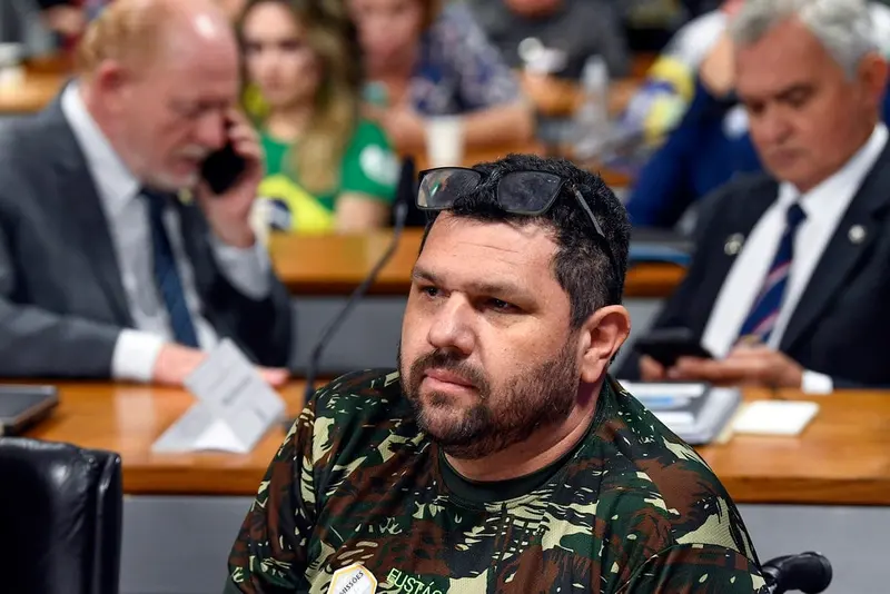 Oswaldo Eustáquio, de barba, camiseta camuflada e óculos escuros na cabeça, sentado à bancada de uma comissão do Senado