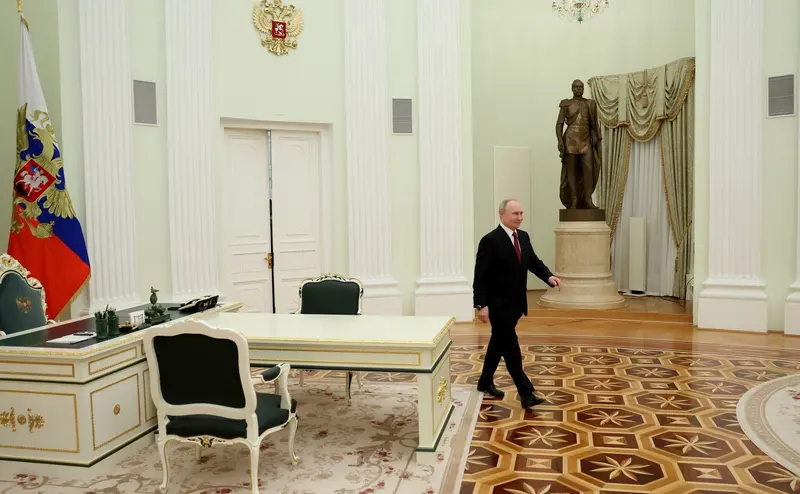 Vladimir Putin, de terno escuro, atravessa a pé um salão do Kremlin de piso de madeira, com uma mesa branca e a bandeira russa ao fundo